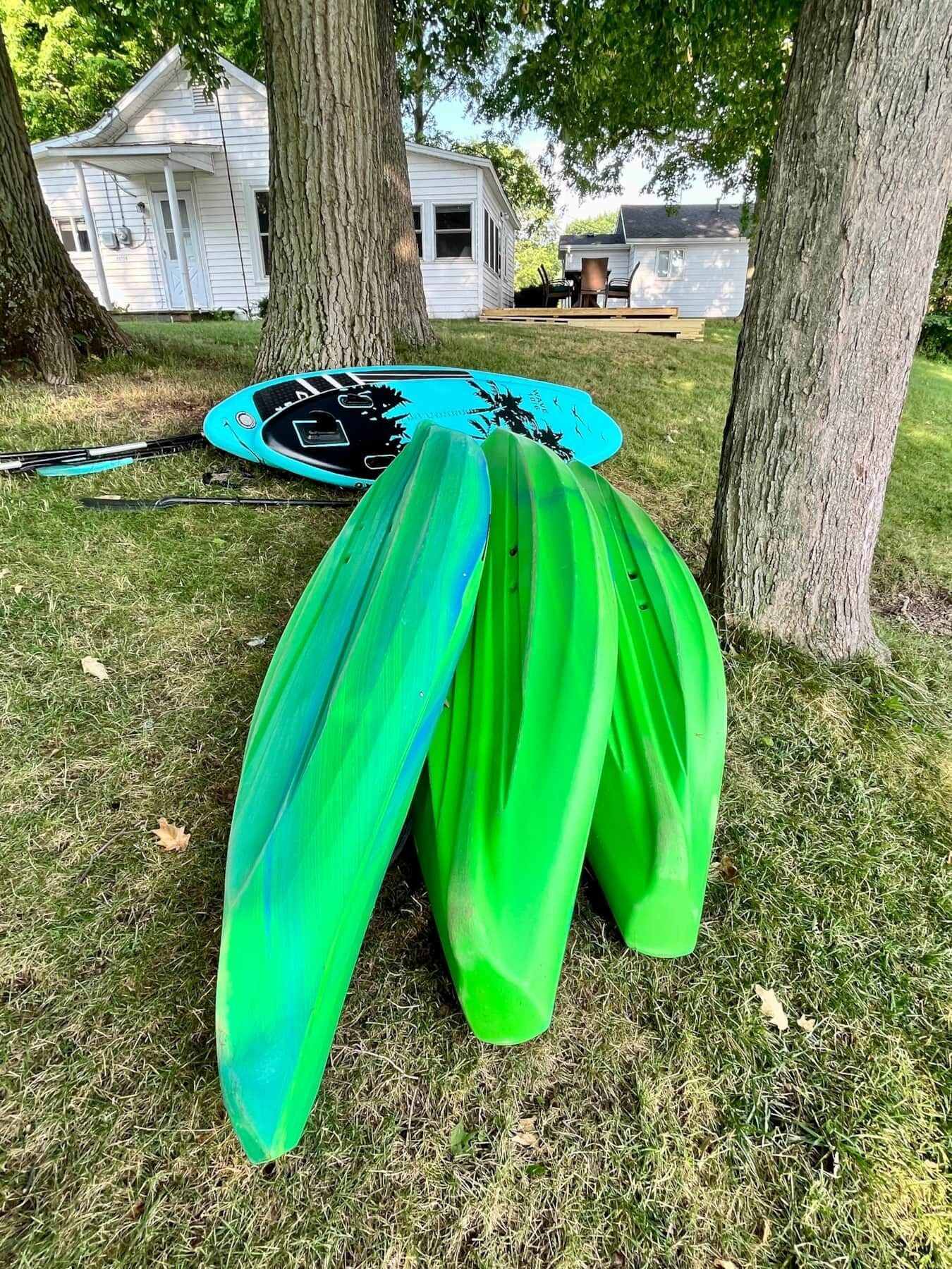 A kayak and paddle leaning against a tree in a grassy area with houses in the background.