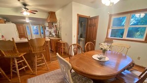 A dining area with a wooden table and chairs, a kitchen in the background, and a window with a view of trees.