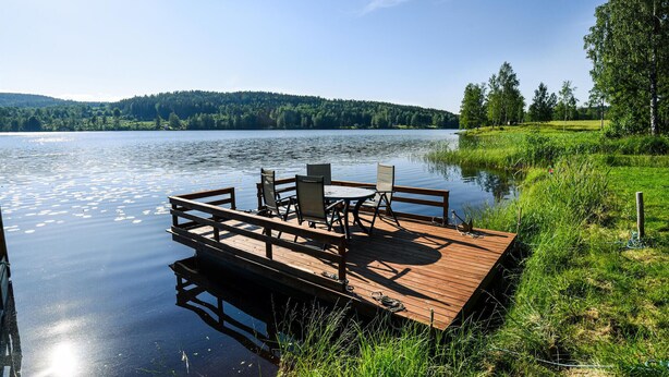 A wooden dock with chairs and a table extending into a calm lake, surrounded by greenery and hills in the background. - 1 bedroom awesome home in åmotfors (Åmotfors)