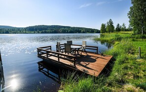 A wooden dock with chairs and a table extending into a calm lake, surrounded by greenery and hills in the background.