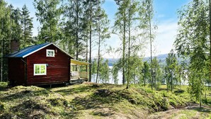 A rustic cabin with a metal roof, surrounded by tall trees and a lake in the background. - 1 bedroom awesome home in åmotfors (Åmotfors)