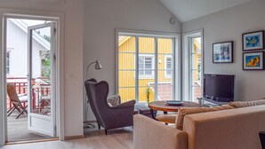 Una sala de estar moderna con un sofá, un sillón y una televisión. Una ventana grande con paredes amarillas se ve desde el exterior, y una puerta da a un área exterior con una mesa y sillas.