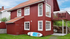 Una casa roja de dos pisos con un balcón, una tabla de surf y una silla en el jardín.