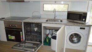 A compact kitchen with a dishwasher, microwave, oven, and a window above the sink. - Casa Modular T2 (Quelfes)