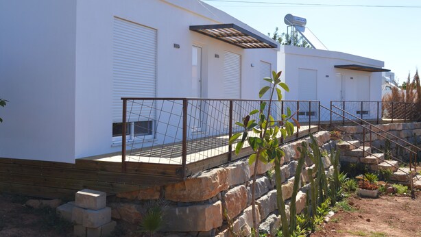 A modern building with white exterior walls, multiple garage doors, and a stone staircase leading to a wooden deck. - Casa Modular T2 (Quelfes)