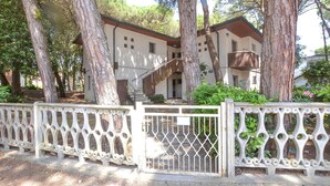 A building with a staircase leading to a second floor, surrounded by trees and a decorative white fence. - Beach front apartment in Rosolina Mare (Rosolina Mare)