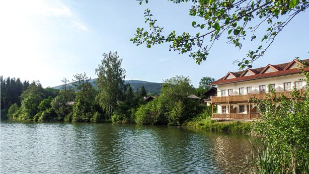 Ett hotell vid en sjö med en träbalkong, omgivet av frodig grönska och med ett berg i bakgrunden.