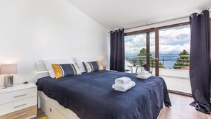 A bedroom with a large bed, a bedside table with a lamp, a view of the outdoors through a large window, and a balcony with a table and chairs. - Awesome Home in Lovran With Wifi and 6 Bedrooms (Lovran)
