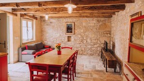 Una habitación rústica con pared de piedra, vigas de madera en el techo, una mesa de comedor de madera con sillas, un sofá con cojines y un banco a lo largo de la pared.