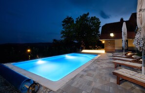 A swimming pool with lounge chairs and umbrellas, surrounded by a stone patio and a building with a sloped roof.