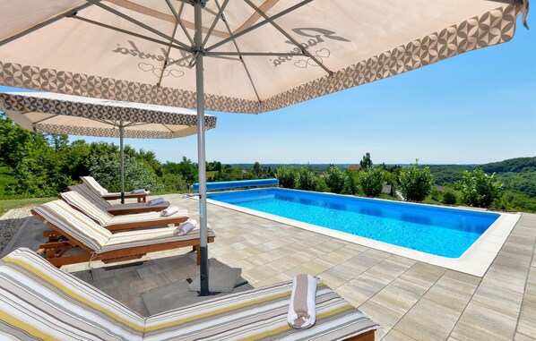 A swimming pool with lounge chairs and an umbrella, surrounded by a tiled deck and greenery.