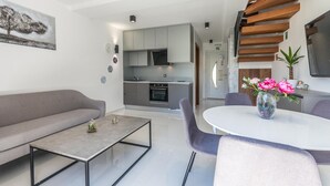 Un saló modern amb un sofà gris, una taula de café de vidre i una zona de dinar amb taula blanca i cadires morades.
