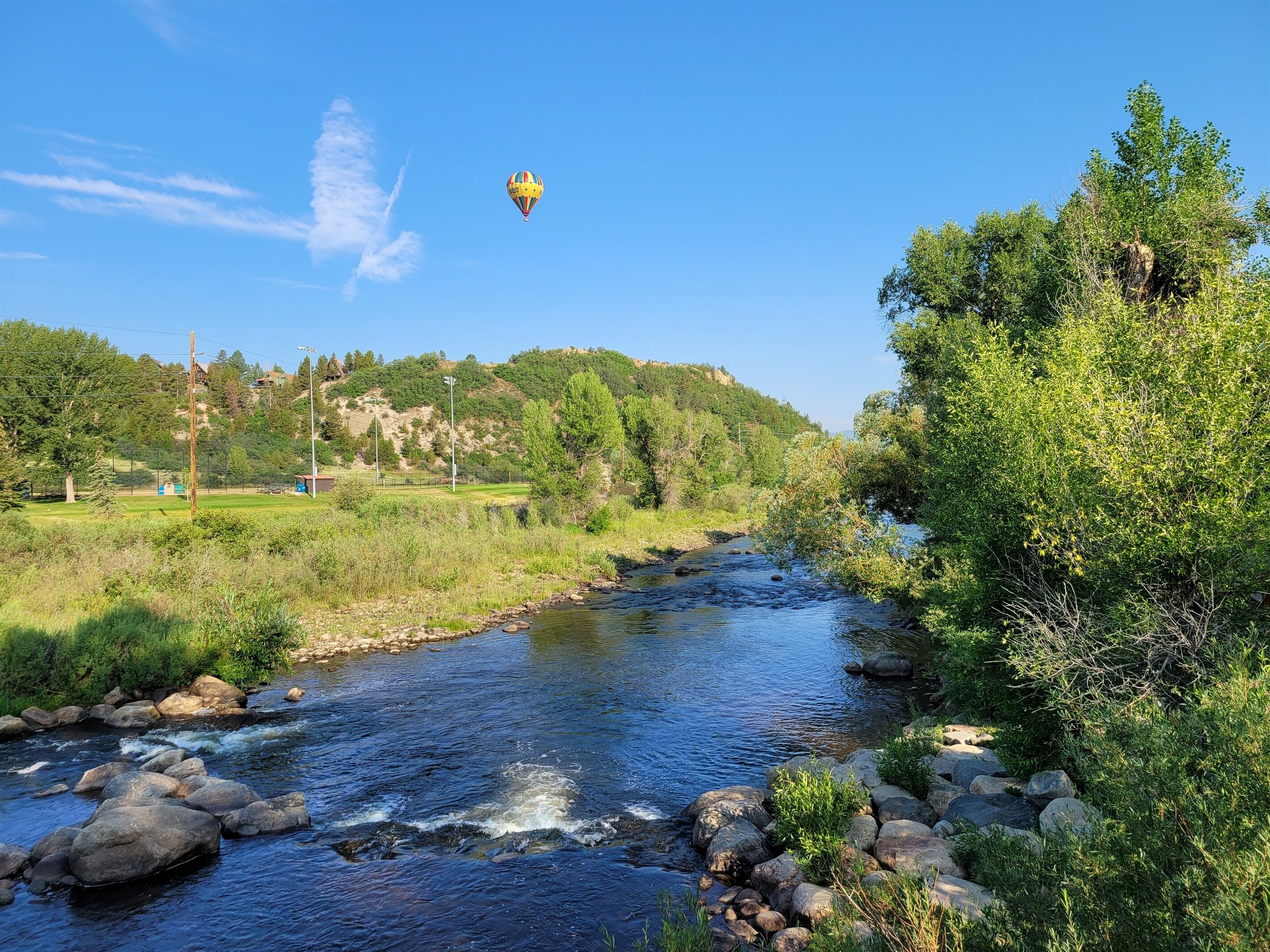 Ein Fluss mit Steinen und Grünpflanzen, ein Heißluftballon am Himmel und eine hügelige Landschaft im Hintergrund.