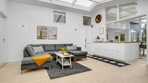 Un salon moderne avec un canapé gris, une table basse et un coin cuisine doté de meubles blancs et d’une fenêtre.
