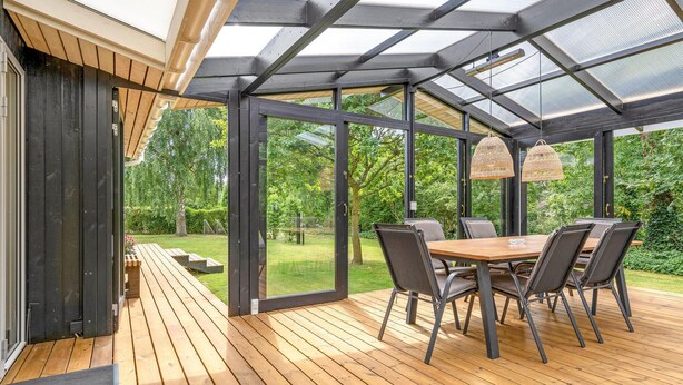 Un patio couvert avec une terrasse en bois, une table et des chaises, et une véranda vitrée offrant une vue sur le jardin.