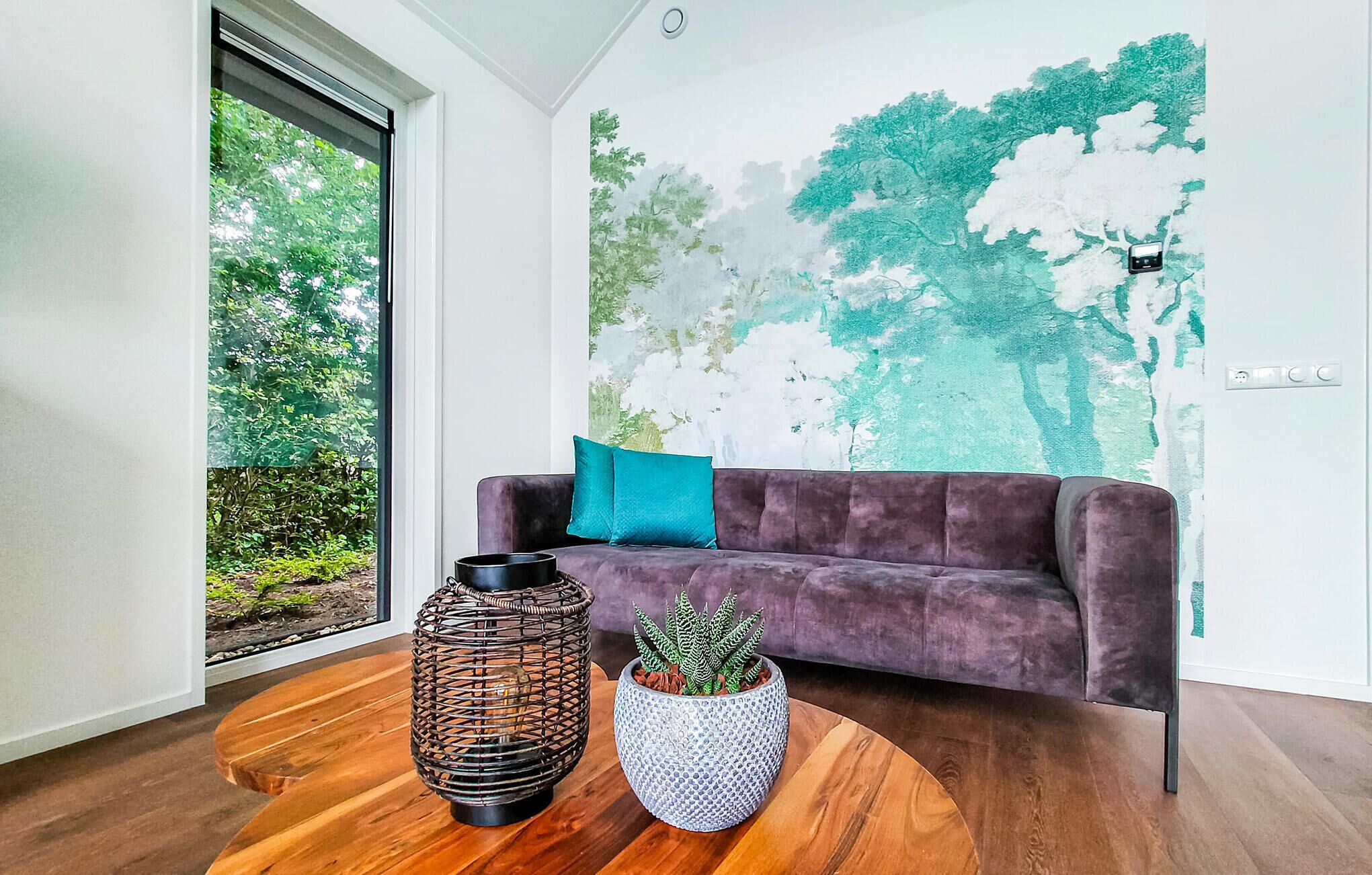Un salon moderne avec un canapé de couleur violet foncé, un coussin turquoise et deux plantes en pot posées sur une table basse en bois.
