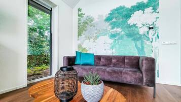 Un salon moderne avec un canapé de couleur violet foncé, un coussin turquoise et deux plantes en pot posées sur une table basse en bois.