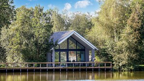 A modern glass house with a metal roof on a wooden dock over a calm lake, surrounded by dense trees.