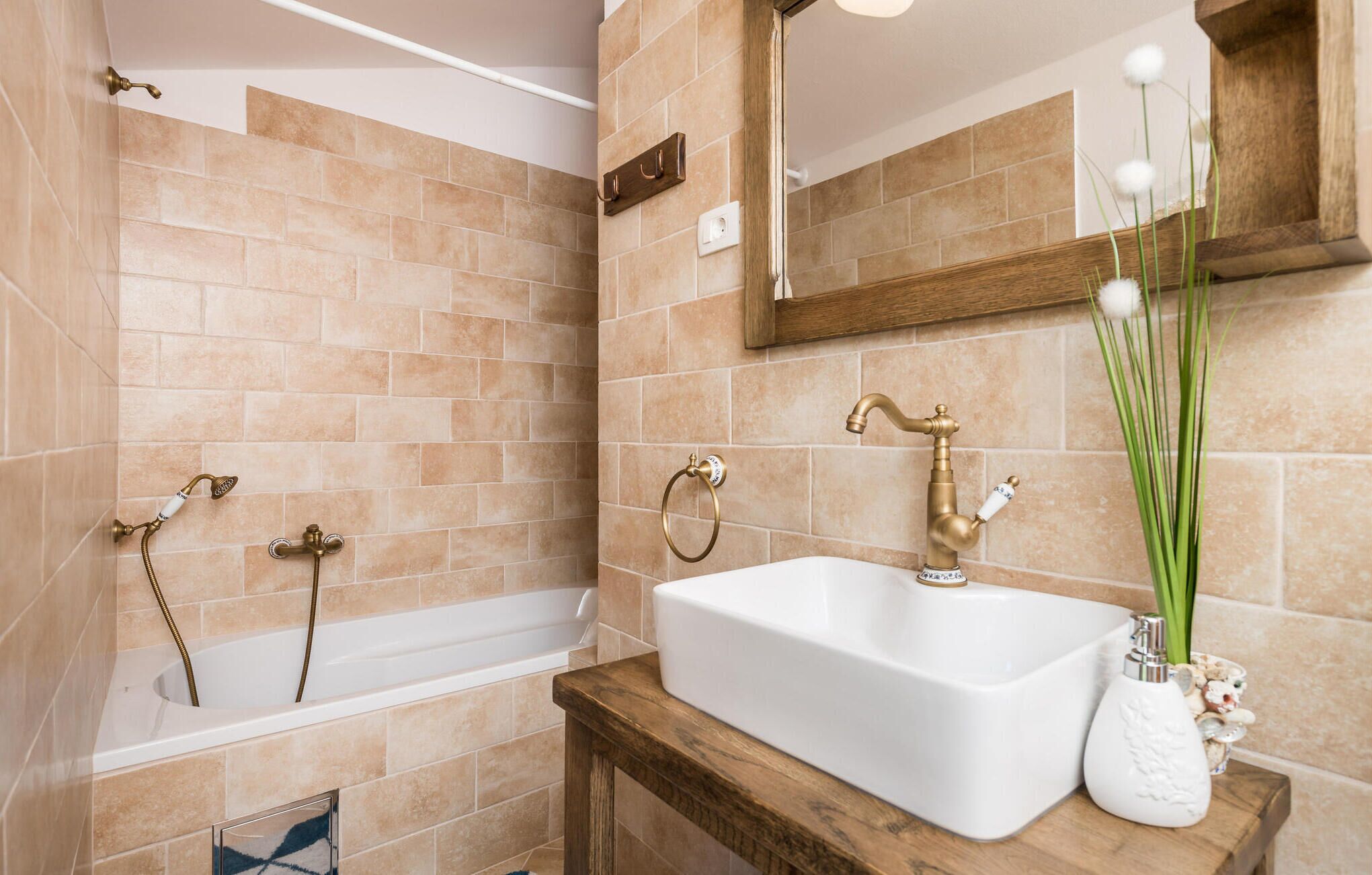 A bathroom with beige tiles, a white bathtub, a wooden vanity, a white sink, and a mirror with a wooden frame.
