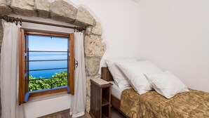 Un dormitorio con pared de piedra, una ventana con cortinas y una cama con almohadas blancas, con vista al mar.