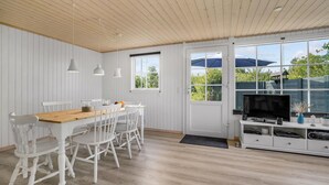 Un coin repas avec une table en bois et des chaises blanches, un téléviseur posé sur un meuble blanc, et de grandes fenêtres donnant sur un paysage verdoyant.