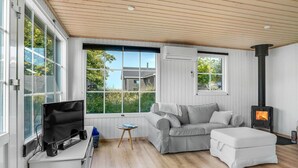 Un salon chaleureux avec un canapé gris, un pouf blanc, un téléviseur posé sur un meuble blanc et un poêle à bois.