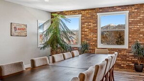 A conference room with a long table and chairs, brick walls, large windows, and potted plants.
