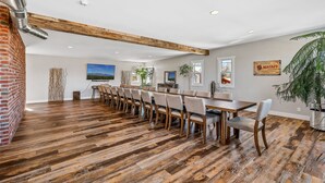 A spacious dining area with a long wooden table and chairs, a brick wall, and a ceiling with exposed beams.