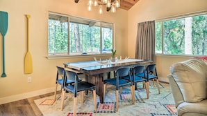 A dining area with a wooden table and blue chairs, large windows with a forest view, and a chandelier. - Pine Mountain Cabin w/ Hot Tub, 2 Mins to Marina! (Groveland)