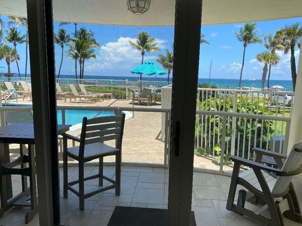 A balcony with a view of a pool and palm trees, a table, and chairs.