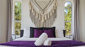Un lit bien fait, recouvert d’une couverture violette et agrémenté de deux serviettes blanches, une décoration murale en macramé et une vue sur les arbres à travers de grandes fenêtres.