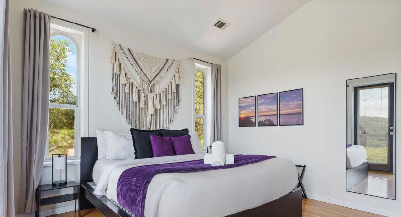 A modern bedroom with a large bed, a bedside table, a mirror, and a view of the outdoors.