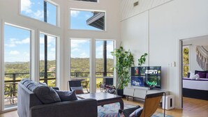 Un salon spacieux doté de grandes fenêtres, d’un canapé confortable, d’un téléviseur à écran plat permettant d’accéder à de nombreux services de diffusion en continu, et d’un balcon offrant une vue sur les collines.