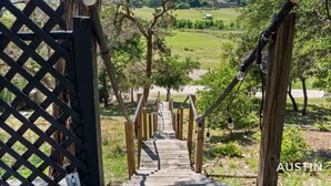 Un escalier en bois mène à un espace verdoyant, parsemé d’arbres, d’où l’on aperçoit une maison au loin.