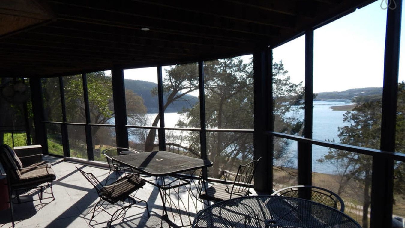Un espacio al aire libre cubierto con una mesa y sillas, grandes ventanales que ofrecen vistas a un lago y árboles.