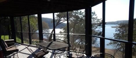 Un espacio al aire libre cubierto con una mesa y sillas, grandes ventanales que ofrecen vistas a un lago y árboles.