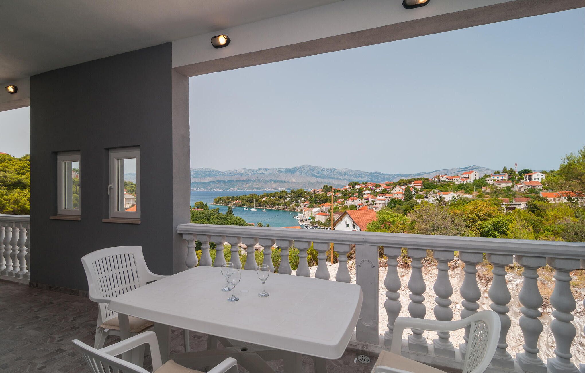 A balcony with a table and chairs, overlooking a coastal town with houses and a view of the sea.