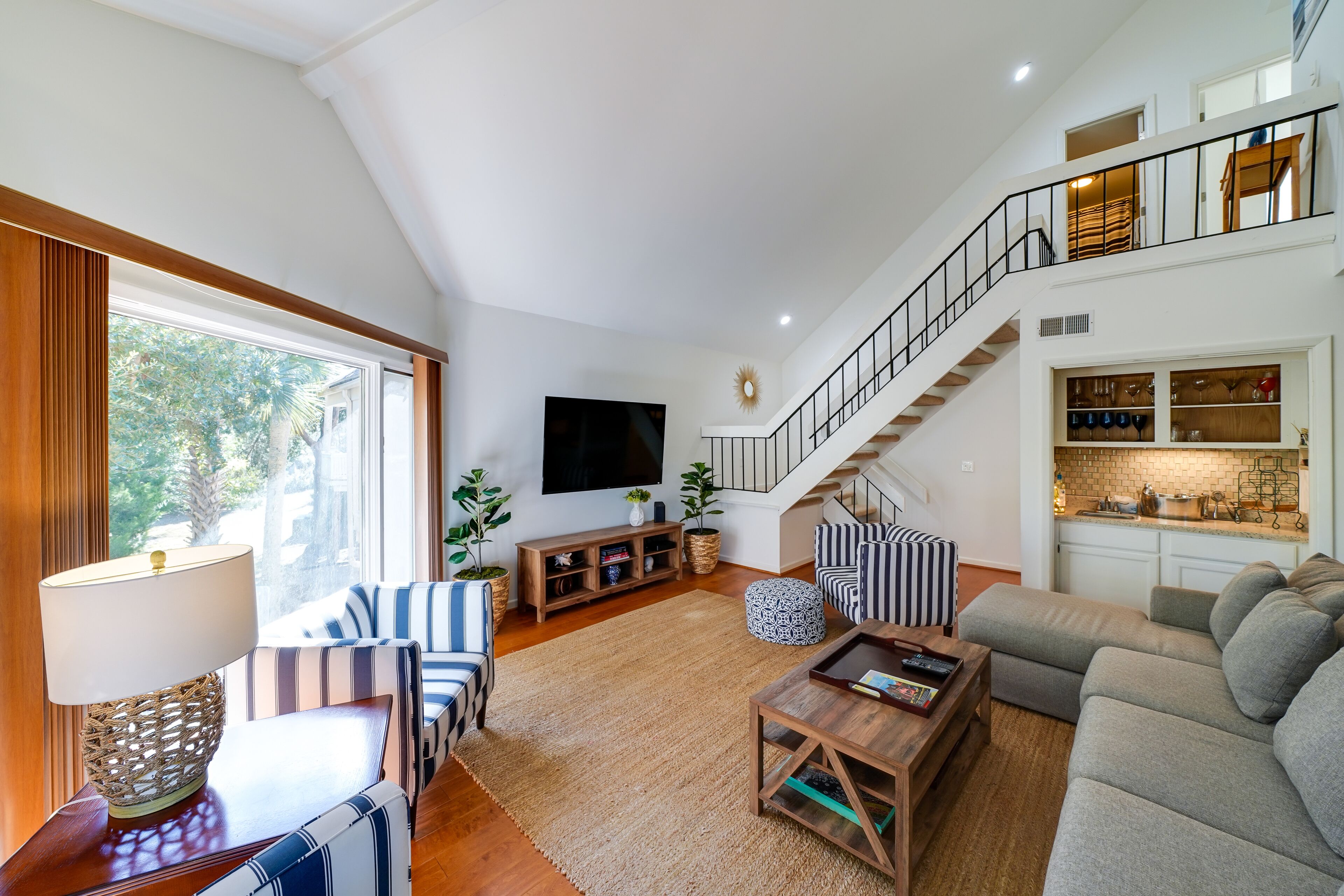 A spacious living area with a high ceiling, a staircase, a flat-screen TV, and a kitchen area with a brick backsplash.