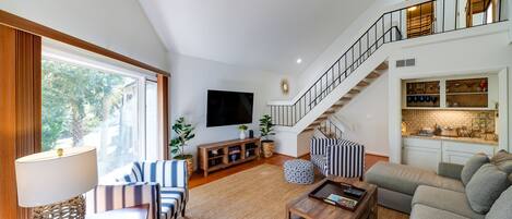 A spacious living area with a high ceiling, a staircase, a flat-screen TV, and a kitchen area with a brick backsplash.