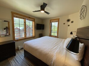 A bedroom with a large bed, a ceiling fan, two windows with wooden frames, a flat-screen TV, and a bedside lamp. - Dakota Lodge 8503 (Keystone)