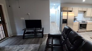 Ein Wohnzimmer mit einem schwarzen Ledersofa, einem Fernseher auf einem Schrank und einer Küche mit hellen Schränken und Geräten.