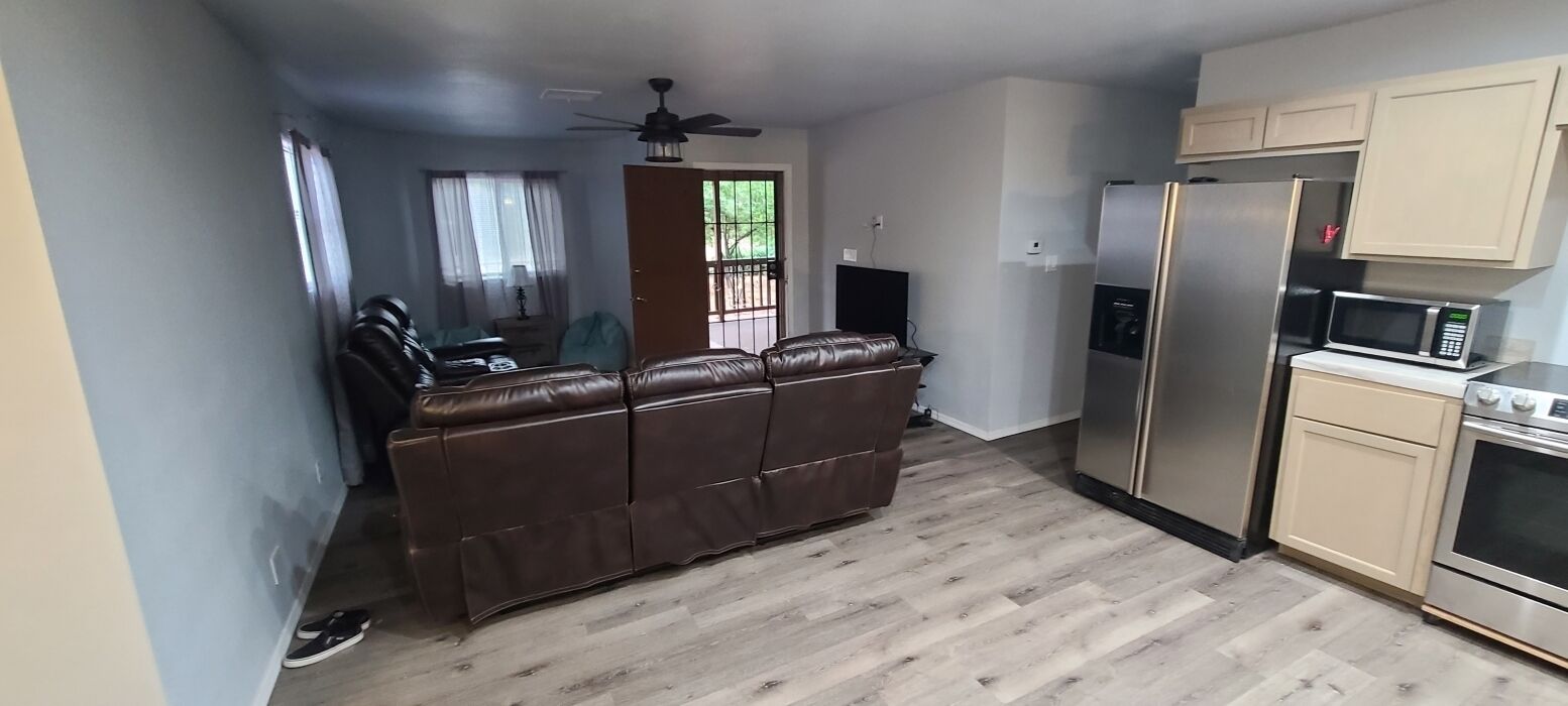 A living room with a brown leather sofa, a ceiling fan, and a kitchen area with a refrigerator and microwave.