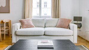 A modern living room with a light grey sofa, two cushions, a coffee table with a book, a wooden chair, a lamp, and a window with curtains. - Modern Brigittenau 1BR w/ AC, Elevator, near Metro U4, by Blueground (Vienna)