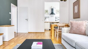 A modern living room with a black coffee table, a grey sofa, and a wooden dining table with chairs. A kitchen is visible in the background with white cabinets and a black range hood. - Modern Brigittenau 1BR w/ AC, Elevator, near Metro U4, by Blueground (Vienna)