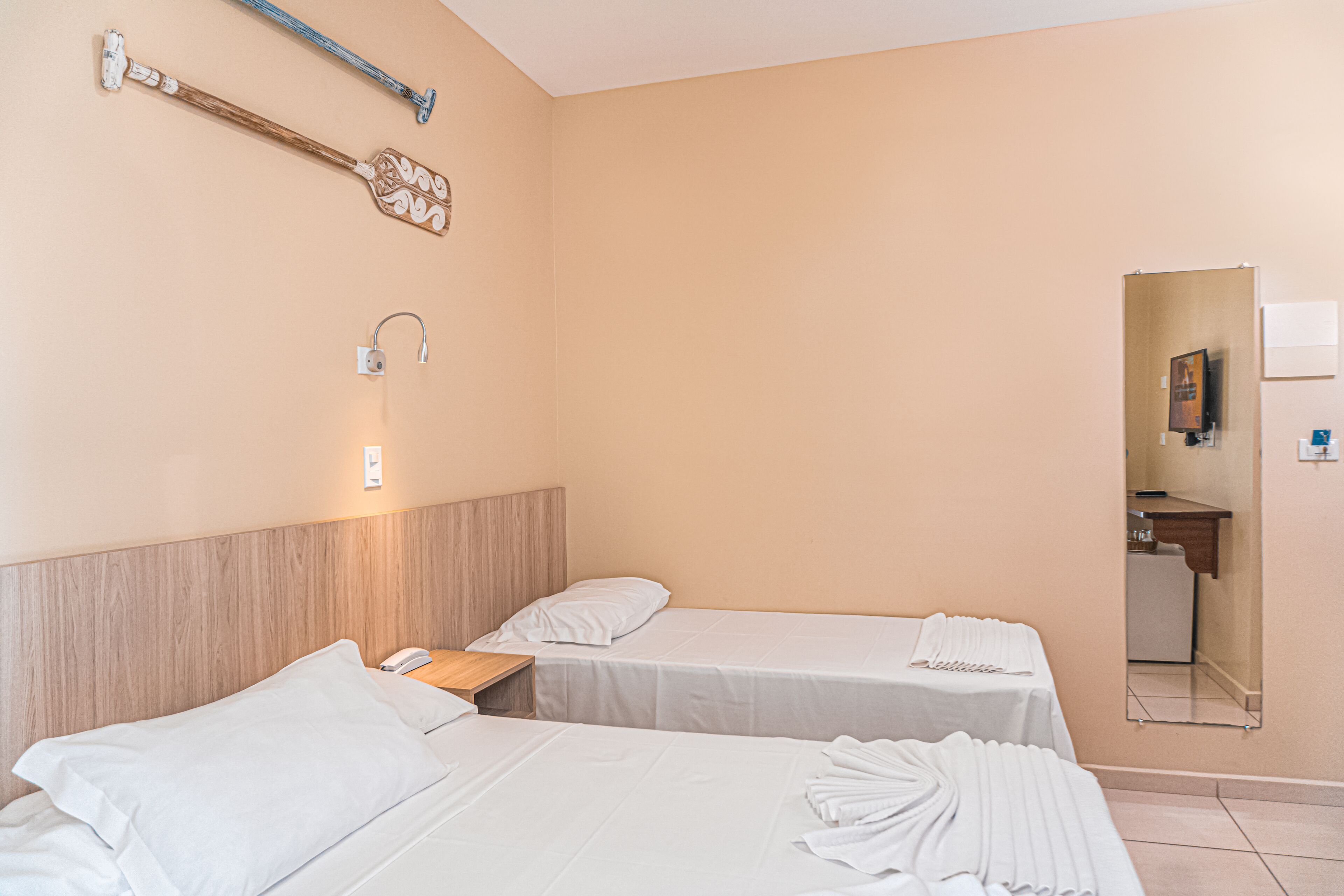 A hotel room with two beds, a wooden headboard, a nightstand, a wall-mounted lamp, a towel rack, and a mirror.