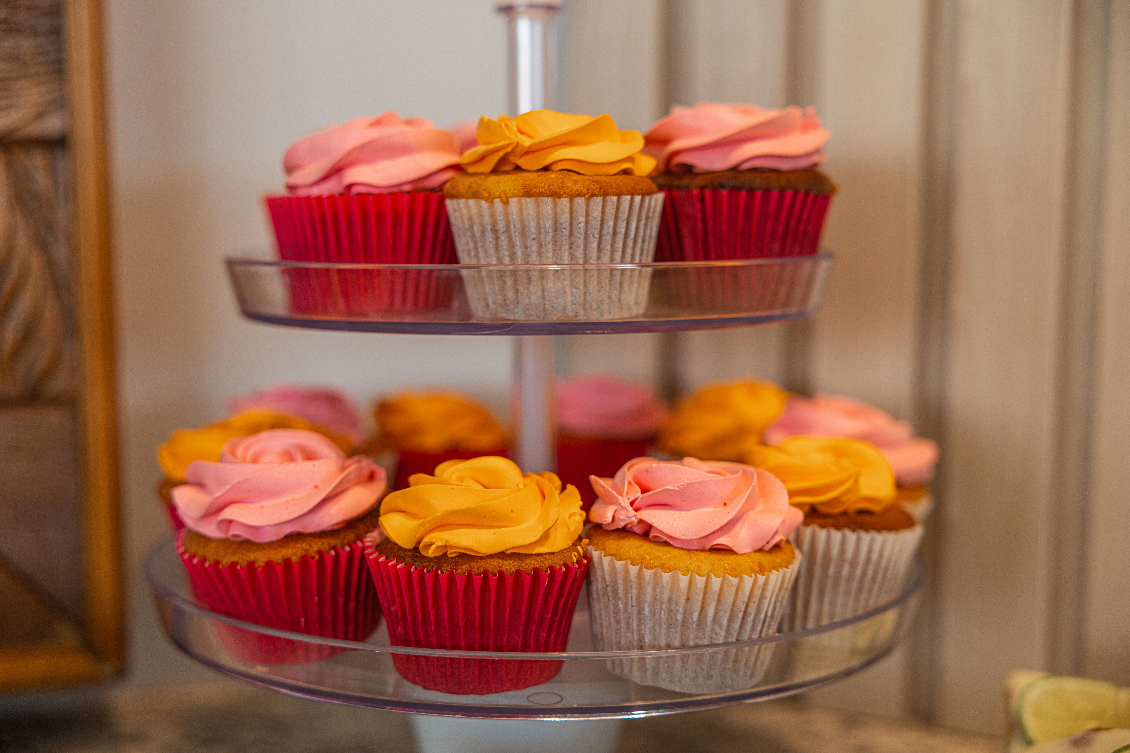 A three-tiered glass stand with cupcakes in red and white paper liners, some with pink and yellow frosting.