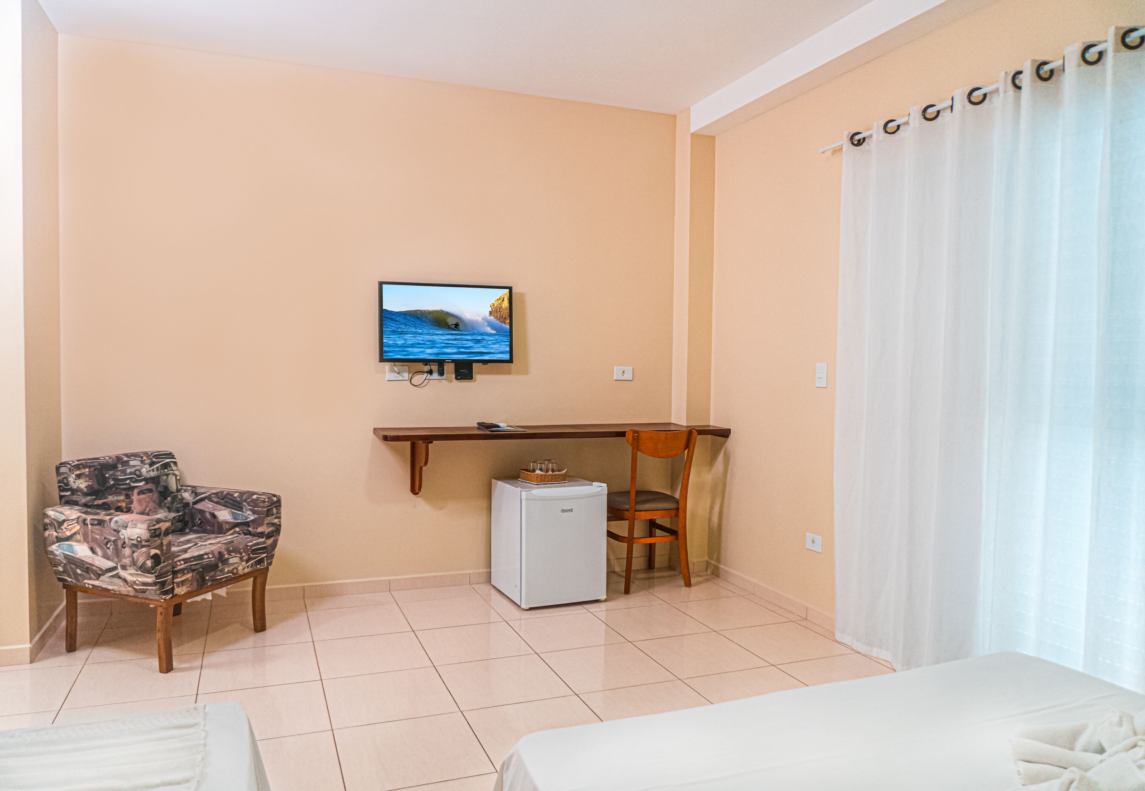 A hotel room with a bed, a desk with a computer, a chair, a small refrigerator, and a television on the wall.