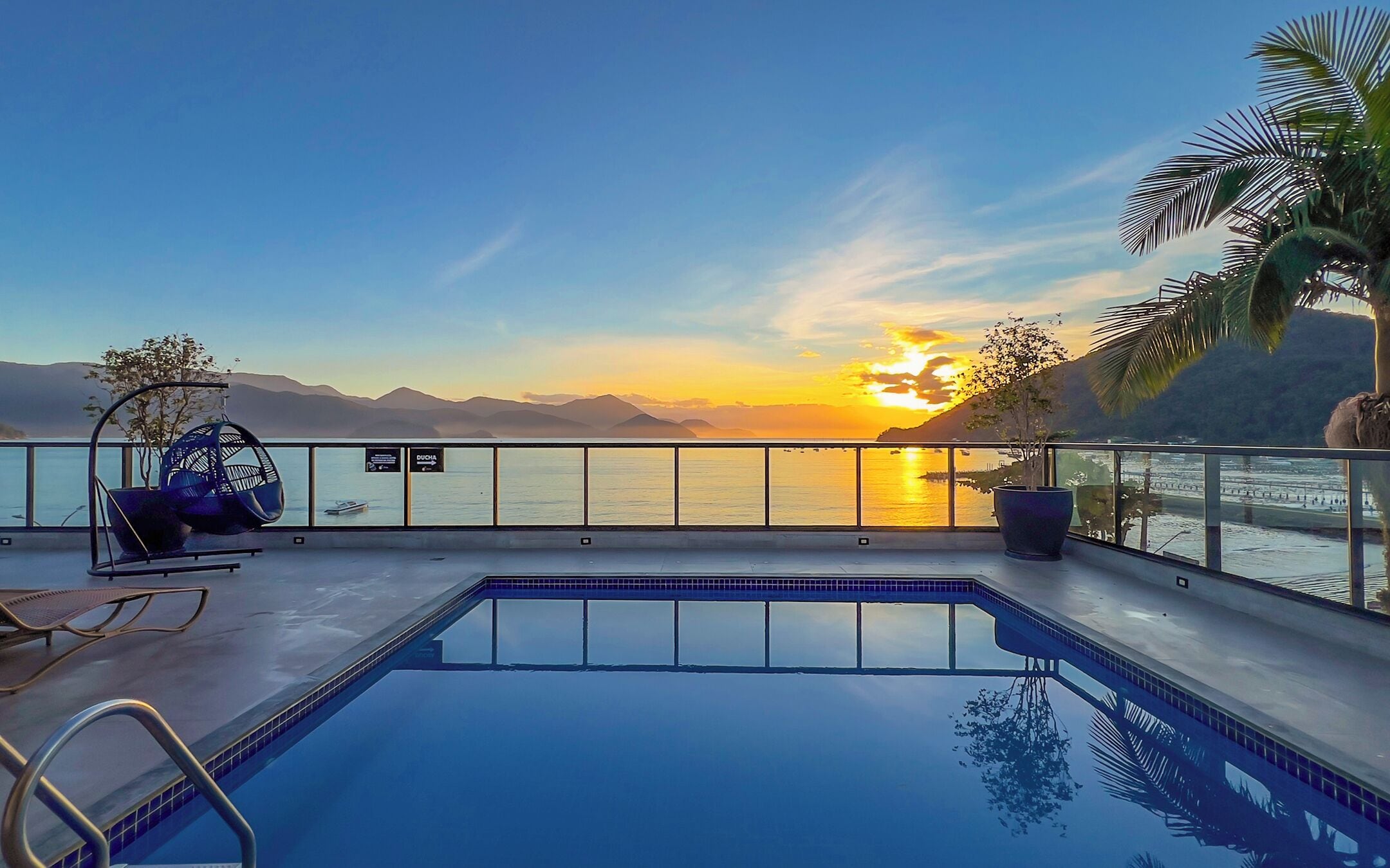 A pool with a view of the sunset over the water, a lounge chair, and a sign for 'DUCHA'.