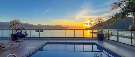 A pool with a view of the sunset over the water, a lounge chair, and a sign for 'DUCHA'.