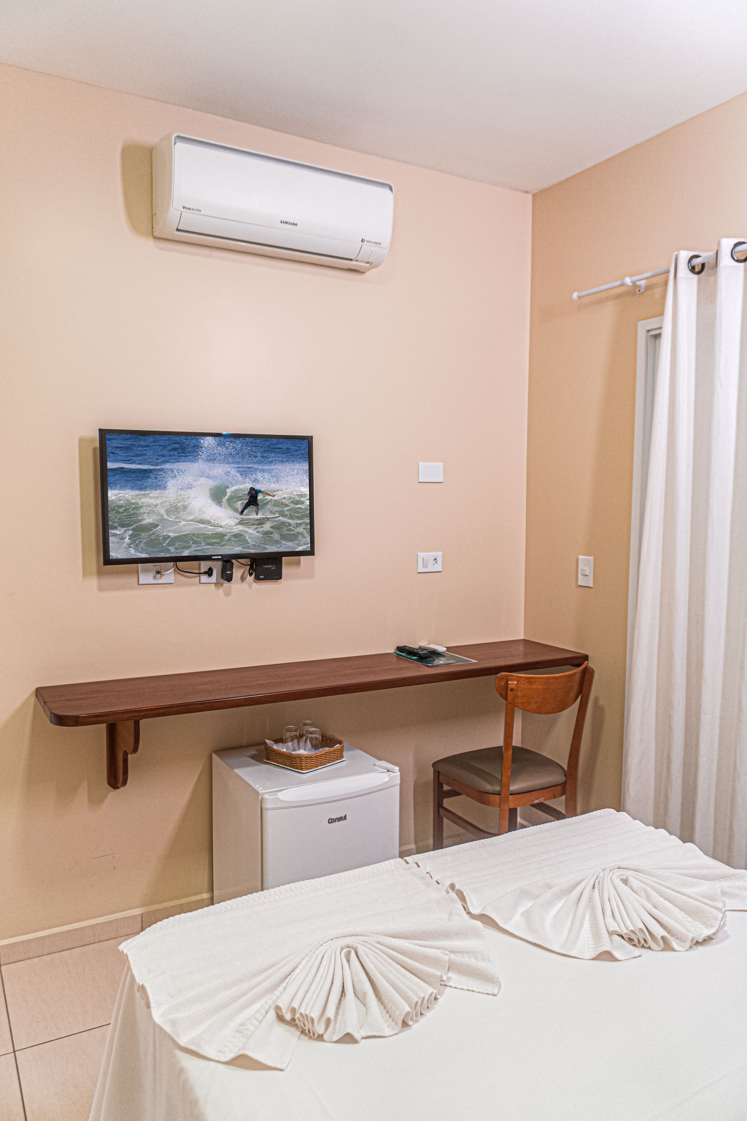 A hotel room with a bed, a desk, a TV, an air conditioner, a small fridge, and a chair.